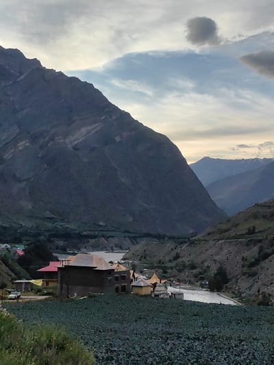 This image depicts a scenic mountainous landscape in Gushal, India, likely captured during the late afternoon or early evening, given the soft, diffused light. The scene is dominated by a massive, dark mountain that slopes dramatically from the upper left corner of the frame downwards, its textured surface suggesting layers of rock. In the mid-ground, a valley unfolds, with a winding river flowing through it, its surface reflecting the muted sky.

Nestled on the slopes overlooking the river are a cluster of buildings, forming a small village or settlement. One prominent building in the foreground features a dark, rough-textured exterior and a multi-layered, somewhat unconventional roof, appearing to be under construction or renovation with scaffolding visible. Nearby, another building sports a bright red roof. A yellow tarp is visible on one of the structures, and a white pickup truck is parked in front of a smaller building. A field of dark, leafy crops, possibly vegetables, occupies the lower foreground of the image, adding a touch of green to the otherwise earthy tones.

Further back in the valley, more structures can be seen, including what appears to be a yellow tent-like structure and a small stupa or monument. The distant mountain ranges are softer in detail and hue, fading into a hazy blue, indicating they are farther away. The sky is partly cloudy, with a mix of grey and lighter tones, suggesting either approaching rain or the transition to dusk. The overall mood is tranquil and serene, highlighting the raw beauty of the natural environment and the humble human presence within it. No visible text is present in the image.