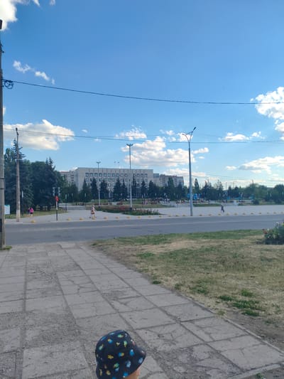 A low-angle, eye-level shot captures a scene in Alchevsk, Ukraine. The foreground features a child wearing a dark hat with colorful cartoon characters. The child is standing on a grey, cracked concrete sidewalk, which extends into the middle ground of the image.

Beyond the sidewalk, a grey asphalt road separates the viewer from a pedestrian area. This area is paved with light-colored tiles and features yellow spherical bollards lining the edge of the road. A traffic light, showing red, is visible on the left side of the road.

The middle ground is occupied by a park-like setting with green grass and mature trees. Several people are walking and milling about in this area, some near flowerbeds. In the background, a large, multi-story building with a light-colored facade and numerous windows dominates the skyline. Power lines and streetlights are also present throughout the scene, and the sky above is a bright blue with scattered white clouds. The overall impression is of a sunny day in a public urban space.