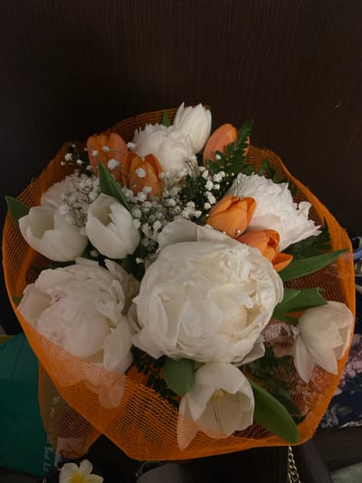 A bouquet of flowers rests on a dark, possibly wooden, surface. The bouquet is wrapped in orange netting. It includes large white peonies, white tulips, and orange tulips. There are also smaller white filler flowers, possibly baby's breath. The flowers are interspersed with green leaves. The overall composition is rich in color and texture. It is not possible to determine the location in the city of Lecco, Italy.