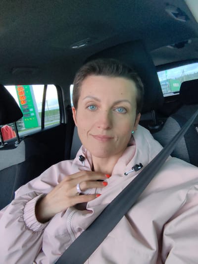 A woman with short brown hair and bright blue eyes is taking a selfie in the passenger seat of a car. She is wearing a pale pink jacket and has her hand on her chest, showing off a light purple ring. A seatbelt is visible across her chest. Through the car window behind her, a green gas station sign with prices displayed in red digits is visible, along with a truck and trees. The scene appears to be at a gas station in or near the city of Fanipalski Sel'skiy Sovet, Belarus.