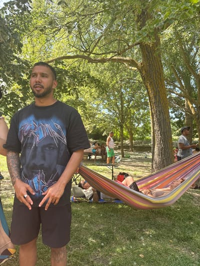In a sun-dappled park with mature trees, a man stands in the foreground, wearing a black t-shirt with a graphic print and black shorts. He has tattoos on his arms and leg and is wearing a ring on his left hand. To his right, a colorful striped hammock is strung between two trees, with a person lying in it, eyes closed. In the background, other people are visible, some seated at a table, others standing. The ground is covered in grass and fallen leaves. The atmosphere suggests a leisurely outdoor gathering. The image does not provide any specific clues to pinpoint its location in Campezo / Kanpezu, Spain.