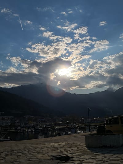 The image captures a bright, sunny day in Skala Potamias, Greece. The sun, partially obscured by clouds, casts rays across the sky and towards a mountain range. A contrail is visible against the blue sky. To the right, a portion of a harbor or waterfront area is visible, with various boats and a few buildings. A yellow van is parked alongside a cobbled or paved walkway.