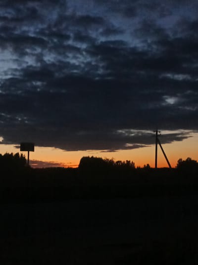 A photograph captures a rural landscape at dusk in the village of Voskresenskoye, Russia. The sky is a dark, moody expanse of clouds, tinged with the faint orange glow of a setting sun near the horizon. Silhouetted against the sky are a line of trees and a power pole with a diagonal brace. To the left, a rectangular sign on a post stands as another dark silhouette. The foreground is entirely black, obscuring any details of the ground or what might be there. The overall impression is one of approaching night in a quiet, remote setting.