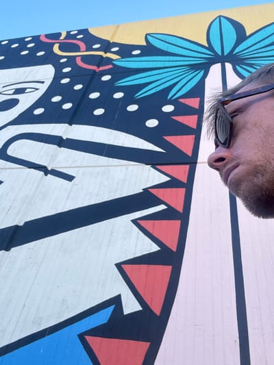 A man wearing sunglasses and a hint of stubble poses in front of a vibrant, modern mural. The artwork features geometric shapes and whimsical designs. Blue leaves accent a yellow background, and a string of colorful squiggles adds a playful touch. Navy paint with white dots meets alternating red triangles. Thick white lines with a black outline resemble sharp mountains, while a blue panel grounds the bottom.