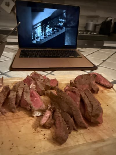 This image depicts an indoor domestic scene in Cava de' Tirreni, Italy. In the foreground, a light-colored wooden cutting board holds numerous slices of cooked meat, characterized by a medium-rare reddish-pink interior and a browned, seared exterior with visible dark seasoning. Behind the cutting board, a rose gold/copper-toned laptop rests open on a light-colored surface featuring square tiles with darker grout lines. The laptop's screen displays a grayscale image of figures on a staircase with a decorative railing. A slender metallic utensil is partially visible on the left, positioned near the laptop. The background, which is out of focus, reveals a kitchen environment, including a dark cooking range, a silver pot situated on the stove, and an additional dark object on a countertop. No individuals are present in the frame.