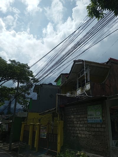 The scene depicts a streetscape in Cibadak, Indonesia, characterized by a mix of residential and commercial structures under a partially cloudy sky. Numerous black utility wires traverse diagonally across the upper and mid-portions of the frame. On the left, a mature tree with dense green foliage stands beside a utility pole. Adjoining the tree, a yellow painted metal fence, topped with barbed wire, encloses a property. A "DIJUAL" (For Sale) sign, white with red lettering, is prominently displayed on this fence. Behind the fence, a two-story building features a yellow lower section and a grey upper section. To the right, another building exhibits a lower facade of exposed, rough-textured brick. A green and white sign on this brick wall advertises "HERBAL SEHAT ROTAN GURAH MATA." The upper level of this building has a yellow exterior with a covered balcony or porch featuring a white railing and a tiled roof structure. A separate white sign for "SOTO AYAM ABANDA" and "MIE & JUICE" is visible on a structure in the background to the far left. No individuals are present in the image.