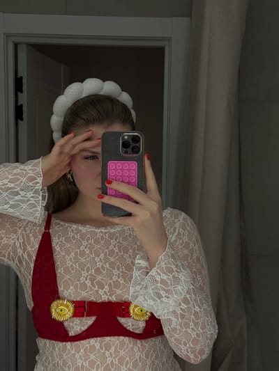 A woman, reflected in a mirror, holds a phone with a pink pop-it grip covering her face. She wears a white, lace, long-sleeved dress with a red, ornamented harness. Her hair is pulled back with a white, frilly headband. The background consists of a room with a white wall and window frame.