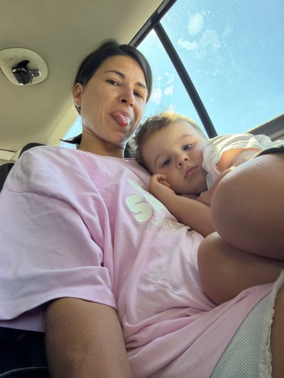 This is a low-angle, close-up selfie taken inside a vehicle, likely a car, as indicated by the presence of a ceiling-mounted latch on the left and a sunroof with a view of the sky above. The image features two individuals: a woman and a young child. The woman is in the foreground, to the left, and is sticking her tongue out at the camera. She has dark hair pulled back and is wearing a light pink oversized t-shirt with a white graphic design. To her right, nestled against her, is a young child, likely a boy, resting his head on her chest. The child has light brown hair and is looking towards the camera with a slightly weary expression. He is wearing a white t-shirt. The overall setting appears to be daytime, with sunlight streaming through the sunroof, creating bright highlights and a slightly hazy atmosphere. The interior of the vehicle is dimly lit, with the exception of the area illuminated by the sunroof. The background is dominated by the sky, which appears blue with some scattered clouds. The scene conveys a sense of intimacy and a candid moment between the two subjects. There is no indication that this image was taken in the city of Kruševica, Montenegro.