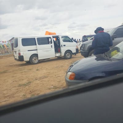 In the city of Wuro Ngangandi, Nigeria, an outdoor scene unfolds under a cloudy sky. A white van, identified by the text "ADAMAWA STATE COMMUNITY AND SOCIAL DEVELOPMENT AGENCY, YOLA (AS-CSDA)," is parked on sandy ground. An orange inflatable structure, possibly a promotional item, is visible on top of the van. A person wearing a dark uniform and a hat stands near the van, facing away from the camera. In the background, several other vehicles, including a gray SUV, and a crowd of people are present. The environment appears to be a rural or undeveloped area, with sparse vegetation and a few structures in the distance. The foreground is partially obscured by a dark-colored car.