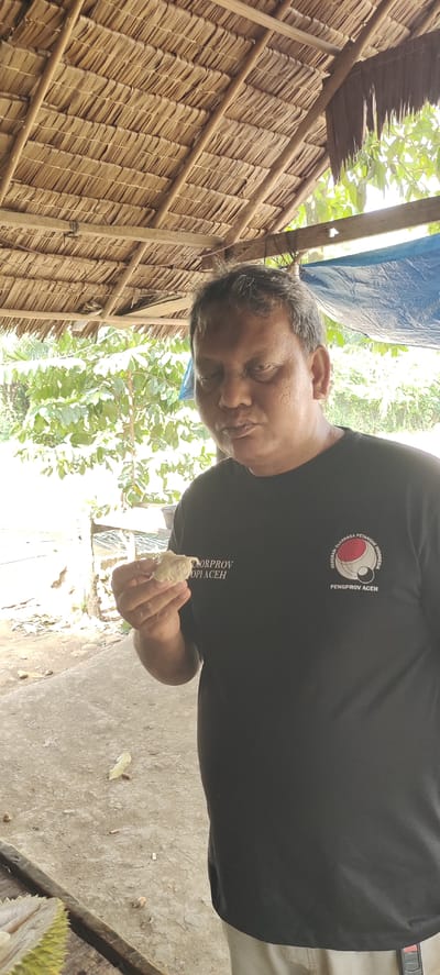 A man with dark skin and graying hair stands outdoors under a thatched roof, holding a piece of fruit to his mouth. He is wearing a black t-shirt with a logo on the left side of his chest and text above it. The text on his shirt reads "DORPROV ACEH" and "PENGPROV ACEH", with the logo appearing to be a circular emblem with red and white elements and a black outline. The man's eyes are closed as he is about to eat. In the foreground, a cut durian fruit is visible on a wooden surface. The background consists of green foliage and a light-colored ground, suggesting a rural or tropical environment. The scene appears to be in Kilometer Delapan, Indonesia.