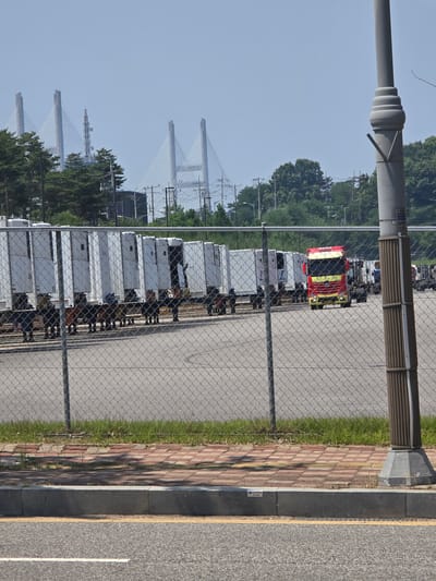The image depicts a street scene in Pyeongtaek-si, South Korea, with a focus on a row of parked white semi-trailers and a red and yellow semi-truck. The semi-truck is positioned in the middle of the frame, facing forward, with its headlights on. The semi-trailers are lined up behind a chain-link fence, and some appear to have their doors open.
In the foreground, a chain-link fence runs horizontally across the image, partially obscuring the view of the semi-trailers and trucks. To the right of the fence is a tall, grey lamppost. Behind the fence, there is a paved area, with some grass and a sidewalk visible between the fence and the road. The road in the foreground is asphalt with a white lane marking.
In the background, several cable-stayed bridges are visible against a pale blue sky. Trees and other foliage are scattered throughout the scene. The overall impression is an industrial or commercial area, possibly a logistics hub or a truck depot. There are no clearly visible people in the image, though it's possible some are obscured by the semi-trailers or fence. The lighting suggests it is daytime, with a bright, hazy sky.