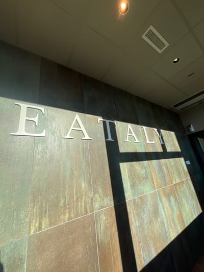The image captures an interior view, likely a hallway or entrance, characterized by a contemporary and clean design. The perspective is an upward diagonal shot, with a tiled floor occupying the right and central portion, and a light-colored wall or ceiling dominating the left side.
Dominating the floor are large, three-dimensional silver or white letters spelling "EATALY". These letters are evenly spaced along the floor, running parallel to the wall on the left. The floor itself is composed of large, rectangular tiles with a textured, variegated appearance, featuring tones of brown, green, and grey, giving it a somewhat distressed or industrial look.
Strong, natural sunlight streams into the space from the right, casting sharp, elongated shadows across the floor and partially onto the letters and the adjacent wall. This indicates a bright, sunny day, likely morning or afternoon when the sun is at an angle. The contrasting interplay of bright light and deep shadows creates a dynamic visual effect.
On the left-hand wall/ceiling, which is a light neutral color (beige or light grey), there are several architectural elements. Two recessed circular lights are visible, one brightly illuminated in the bottom left, and another further up. A rectangular air vent or grille is also prominently featured on this surface. A dark doorway or frame is partially visible in the upper right corner, adding depth to the scene. No people are present in the image.
The visible text "EATALY" clearly indicates the location is associated with the Eataly brand, an Italian marketplace chain. Given the provided location context of Dorno, Italy, this image likely depicts an Eataly establishment in or around that area.