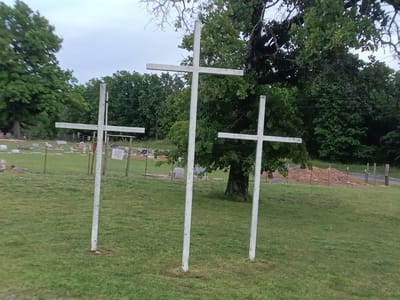 The image depicts three white crosses standing on a grassy lawn. Behind the crosses, there is a cemetery with tombstones and a variety of trees. The area appears to be outdoors, possibly on a cloudy day. It is identified as being located in Stilwell, Oklahoma, United States.