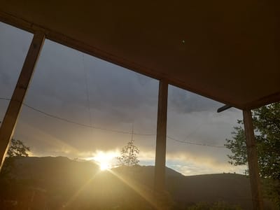 The image captures a dramatic sunset viewed from a covered porch or overhang, likely in Areni, Armenia. The foreground is dominated by the wooden support beams of the structure, framing the expansive landscape beyond.

The sky is a mixture of dark, stormy clouds and areas of bright, golden light. The sun, a brilliant, lens-flared orb, is low on the horizon, casting long, intense rays that pierce through the clouds and illuminate the rolling hills and mountains in the distance. The sunlight creates a strong contrast between the illuminated and shadowed parts of the scene, giving it a dynamic and almost ethereal quality.

A few trees are visible, silhouetted against the bright sky or catching the last rays of light. One small, spindly tree stands prominently in the middle ground, its branches reaching upwards. Wires are strung across the view, suggesting the presence of civilization nearby, perhaps a rural dwelling.

The overall mood is one of serene beauty, albeit with a hint of impending weather due to the dark clouds. The intense sunlight and the natural landscape evoke a sense of awe and tranquility. The time of day is clearly sunset. No people or text are visible in the image.