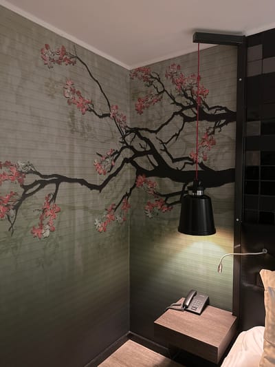 The image depicts a hotel room interior. A black wall-mounted lamp is visible, with its cord running across the wall. The wall features a wallpaper with a pattern resembling a black tree with pink blossoms against a striped background. A telephone is placed on a wooden bedside table. There is a small flexible reading light fixed to the same table. The location is likely in Genk, Belgium, but that cannot be verified from the image itself. No people are visible.