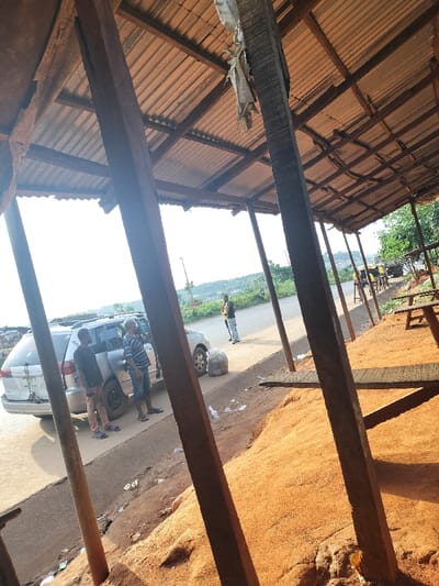 The scene is set outdoors in Oluku, Nigeria, viewed from within a partially enclosed structure. In the immediate foreground, three prominent dark wooden beams frame the composition, two on the left and one on the right. Overhead, a corrugated metal roof, supported by visible wooden rafters and beams, extends across the frame. Below the roof on the right, a large mound of reddish-orange material, consistent with sawdust or fine earth, is present, with crude wooden planks serving as benches or tables.
A paved road occupies the midground. A silver minivan or SUV is parked on the roadside, facing left, with its passenger-side sliding door partially open. Two males stand next to the vehicle; one in a dark shirt and light pants is near the rear passenger door, while another in a striped short-sleeved shirt and blue shorts is closer to the open front passenger door. A gray bundled object, possibly a trash bag, rests on the ground near the minivan's front wheel. Litter is scattered on the ground near the roadside. Further down the road, a third male in a yellow shirt and dark pants stands alone, facing away from the camera.
The background reveals a landscape of rolling hills under a bright sky, suggesting daylight conditions. Utility poles are visible along the road. To the far right, a large vehicle, possibly a truck, is parked or moving slowly, with several indistinct individuals nearby, some appearing to be on bicycles or motorcycles.
