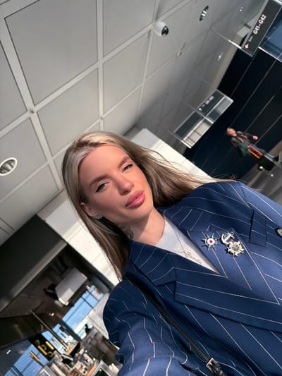 The image captures an upward-tilted selfie of a blonde woman located in a modern airport terminal. The woman, positioned in the foreground, has long blonde hair, prominent lips, and visible eyeliner, with a small tattoo present on her left neck. She wears a navy blue pinstriped blazer over a white ribbed top and a silver necklace. The blazer is embellished with three distinct pins: a red and white star-like emblem, a Russian naval flag-like emblem, and a multi-pointed star-shaped medal. A white dome security camera is mounted on the paneled ceiling overhead. In the mid-ground, a black sign with white lettering explicitly displays "Gate G41-G42." A blurry figure of another individual pulling luggage is visible in the distant background. The location is identified as Munich Airport (MUC), which is situated in Freising, Germany.