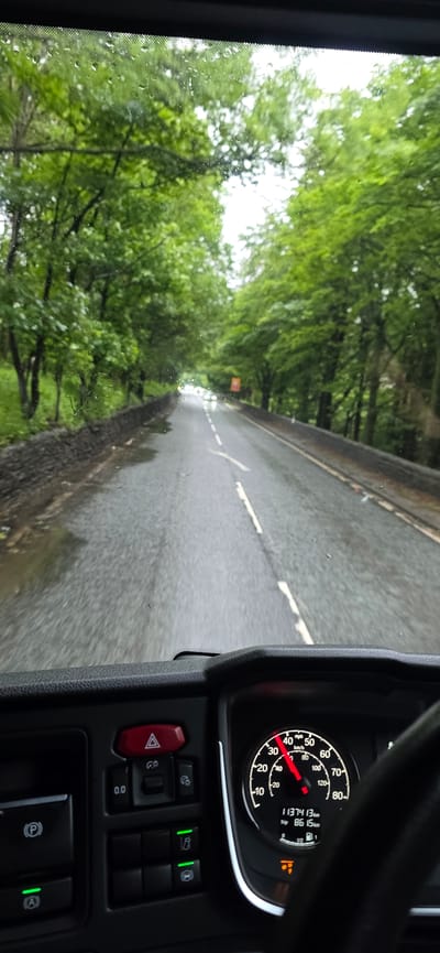 The image is taken from the interior of a vehicle, looking forward through the windshield. The windshield has raindrops on it, indicating wet weather. The road ahead is wet and appears to be a two-lane road lined with trees on both sides. There is a stone wall on the left side of the road. The scene suggests a rural or semi-rural setting.
The dashboard of the vehicle is visible in the foreground, with various buttons and controls. A speedometer is also visible, showing the vehicle is traveling at approximately 40 mph. The speedometer also displays trip information, indicating 11374.13 km have been traveled and the current trip is 86.15 km.
The location is identified as the city of High Peak, United Kingdom. This suggests the image was captured on a road within this region. The overall atmosphere is one of driving in damp, green, and possibly forested surroundings.