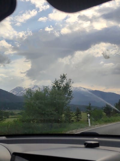 View from inside a car driving on a road. The sky is cloudy. Mountains with snow-capped peaks are visible in the distance. Green trees and vegetation are present along the roadside. A road marker stands on the right side of the road. Location is potentially near Dobrinishte, Bulgaria based on the presence of mountain ranges.