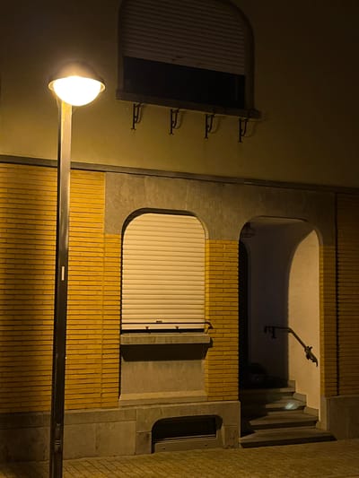 The image is a night-time outdoor shot of the exterior of a building in Saint-Vith, Belgium. The building features yellow brick walls and concrete accents. A street lamp with a bright white light is prominent on the left side of the frame. The building has a large, arched window covered by a beige roller shutter. To the right of the shuttered window, there is an arched doorway with a set of stairs leading up into the dark interior. A metal handrail is attached to the wall beside the stairs. The ground in front of the building is paved. There are no people or animals visible in the scene.