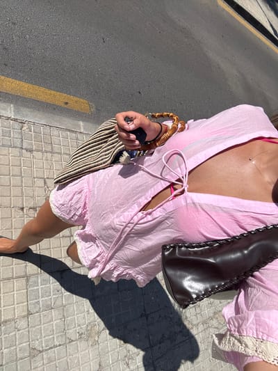 An overhead view captures an individual standing or walking on a tiled sidewalk adjacent to an asphalt road in Cas Català-ses Illetes, Spain. The person, partially visible from the chest down, wears a light pink, loose-fitting, crinkled cover-up or dress with a tied neckline and white lace trim along the hem, revealing a bright pink bikini top underneath. Their tanned right arm extends forward, with the hand grasping a black object, possibly a smartphone or vehicle remote, along with a set of keys. Adorning the right wrist are several bracelets, including one with wooden, bamboo-like beads and a thin black band. Held in the right hand is a striped bag, possibly woven straw, with a distinct bamboo handle. A dark brown, leather-like bag with decorative braided stitching rests on the left side of the person's body. The ground comprises light grey square-patterned sidewalk tiles on the left and dark grey asphalt on the right, featuring a double yellow line painted along its edge. Strong, defined shadows of the person and their belongings are cast onto the sun-drenched sidewalk, indicating clear, bright daytime conditions.