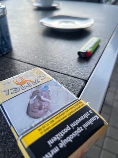 A close-up, overhead shot shows a pack of Camel cigarettes placed on a dark, textured outdoor table. The cigarette pack is yellow and features the Camel logo with a camel graphic. The lid of the pack is partially open, revealing a health warning image of a person's face with medical tubing attached. To the right of the cigarette pack, a green and red lighter lies on the table. In the background, out of focus, a white coffee cup and saucer are visible on the table. A portion of a blue Eiffel Tower-shaped salt or pepper shaker is visible on the left side of the image. The scene appears to be set at an outdoor seating area of a cafe or restaurant. The image has a slightly blurred quality, suggesting it was taken with a mobile phone camera.