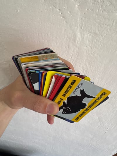 A close-up shot shows a hand holding a stack of plastic cards against a textured white wall. The top card is a yellow and gray Killfish Discount Bar Payment Card featuring a black fish, possibly a piranha. Text on the card includes "KILLFISH DISCOUNT BAR PAYMENT CARD," "0221612," "WWW.KILLFISH.RU," and "8 (800)." Other cards in the stack are multicolored and partially visible. Text on the other cards include "ROSAN 128891 05/11" and "105 500.55.50."