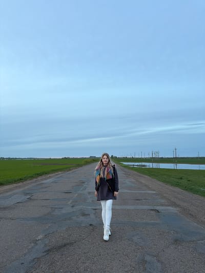 A woman stands on an asphalt road in Azyakul', Russia. The road is adjacent to green fields. Overhead power lines are visible. The sky is overcast. The woman wears white pants, a dark jacket, and sneakers. She has long blonde hair.