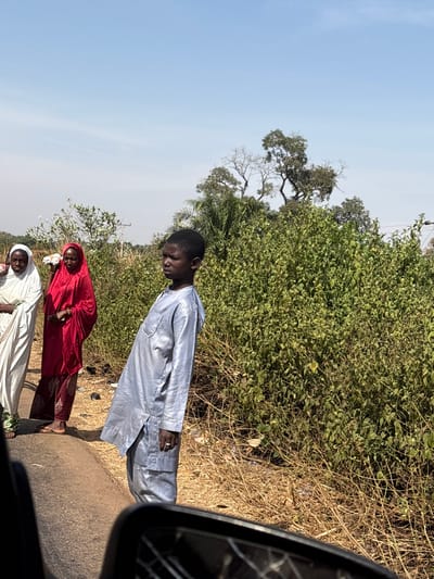 The image depicts a roadside scene in Nandu, Nigeria, during daylight hours under a clear blue sky. In the foreground, viewed from a low angle, a cracked vehicle side mirror is partially visible in the lower right.
The central subject is a young male, likely a pre-teen, standing in profile facing left. He wears a light grey or silver long-sleeved tunic and matching trousers, with a ring on his right ring finger. He stands on the dirt shoulder adjacent to a dark paved road.
To the left and slightly behind him, two adult females are positioned on the road's edge. The leftmost woman wears a white robe and white head covering. The woman next to her wears a red robe and red head covering, and holds multiple white, cylindrical objects in her right hand, resting near her chest. Both women appear to be barefoot.
The background features dense green foliage and scrub immediately behind the individuals, extending into the mid-ground on the right. Further back, taller trees, some with sparse foliage, are visible against the sky, along with a palm tree on the far left. The ground on the roadside not covered by lush vegetation consists of dry, light brown soil and sparse dead grass.