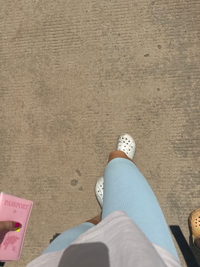 The image shows a person's legs and feet standing on a concrete surface, possibly a sidewalk or path. The person is wearing light blue ribbed leggings and white crocs. They are holding a pink passport in their left hand, with a neon yellow sticker on it. A second person's foot wearing a tan croc is visible on the right side of the image. The concrete surface has visible grooves and is textured. The scene suggests an outdoor urban or pedestrian environment. The location is not specified as Xayden, Laos.
