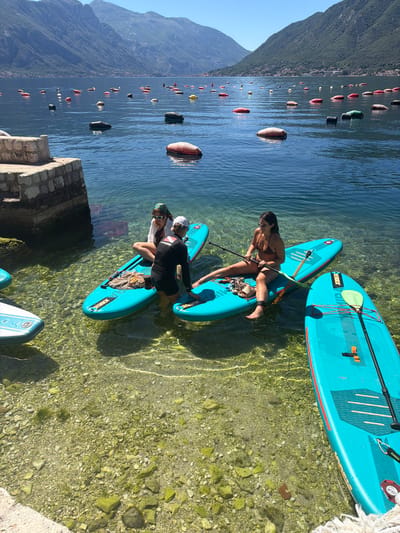 The image depicts a scene on a clear, sunny day in Dražin Vrt, Montenegro. In the foreground, three individuals are preparing to paddleboard in shallow, clear water with a rocky bottom. The water is transparent, revealing the details of the seabed.
A woman in a brown bikini stands on a blue paddleboard, adjusting a paddle. Two other people, one wearing a white hat and another with sunglasses, are on a second blue paddleboard. They appear to be communicating.
There are four blue paddleboards in the water, with one lying upside down. The paddleboards have a brand logo: “JOBE”.
In the background, numerous buoys of various colors (red, black, yellow, and striped) float on the water's surface. A steep, green mountain rises sharply from the water’s edge on the left side of the frame. A stone structure or dock is partially visible in the lower left corner.