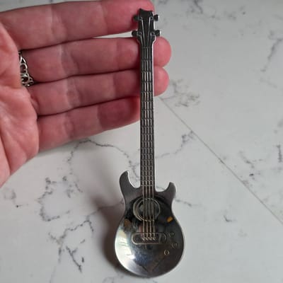 In Beaverton, Oregon, a hand with a decorative ring is holding a silver spoon shaped like a guitar. The spoon has a detailed design, including a headstock with tuning pegs, a fretted neck, a body with a soundhole and strings, and control knobs. The background is a white marble surface.