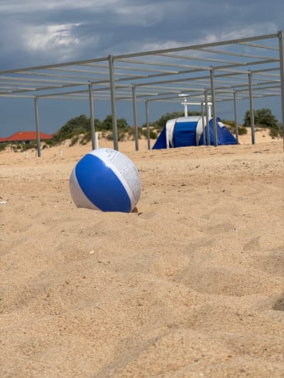 In Vityazevo, Russia, a blue and white inflatable ball lies on a sandy beach. Behind it, a blue and white tent is set up, partially obscured by a metal frame structure. In the background, a red-roofed building and green foliage are visible against a cloudy sky. The sand is textured with small pebbles and some scattered debris. The scene appears to be a beach campground or a recreational area.