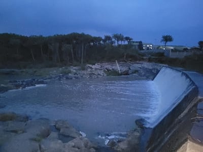 The image portrays a twilight or nighttime scene in Kinanie ward, Kenya, centered on a body of water flowing over a concrete weir. The water, appearing murky grey-blue with visible ripples and some foaming, cascades over a curved concrete spillway from left to right. The left bank is composed of numerous rocks and sparse vegetation, backed by a dense line of tall, slender, dark trees. The right bank primarily consists of rocky terrain and sparse foliage, with a light-colored pole partially visible among the rocks. In the distant background on the right, a multi-story building with several illuminated windows is visible, suggesting an active structure. A concrete block wall or fence is also present further right. The sky is a deep, dark blue, indicative of low light conditions. No individuals, animals, or explicit actions are observed within the frame.