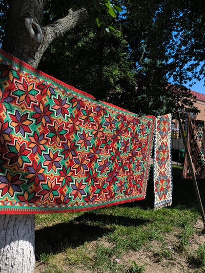 Multiple vibrantly patterned textiles are displayed outdoors under bright sunlight in Illintsi, Ukraine. In the foreground, a large, rectangular textile, likely a carpet or tapestry, featuring repetitive geometric star-like motifs in red, green, orange, purple, pink, and black, is stretched horizontally. Its left end is anchored to a textured, light-colored tree trunk. To the right, a narrower, vertically hanging textile with blue, white, red, and yellow patterns and fringed edges is visible. Further to the right, additional textiles are partially visible, suspended vertically from what appear to be wooden poles. The ground consists of green grass with small patches of dirt and sparse small plants, with distinct shadows cast. The background features dense dark green foliage of trees, a clear blue sky, and a building with a reddish-orange tiled roof. No individuals are visible in the scene.
