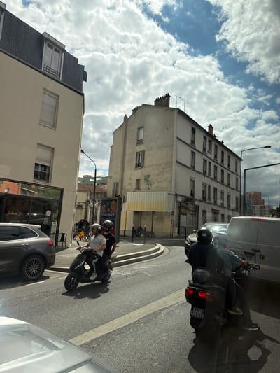 The image depicts a street scene in Alfortville, France, on a cloudy day. The main subjects are three people on a scooter, two of whom are in the foreground. The person in the front is wearing a dark helmet and a dark jacket, and the person behind them is wearing a white shirt and a helmet. A third person, mostly obscured, is behind them. They appear to be riding or stopped on the scooter.
The setting is an urban environment with buildings lining the street. On the left, there's a light-colored building with multiple windows and a modern, dark-tiled roof on an upper section. Further back, a taller, older building with a grid of windows dominates the background. The street itself is paved asphalt, with a concrete curb and a painted line indicating a lane.
Notable details include the scooter's license plate, which is partially visible and appears to read "FK 171-EQ". Shadows are cast on the ground, suggesting the sun is present, albeit partially obscured by clouds. The overall mood seems casual, possibly depicting friends or individuals commuting.
There are also glimpses of other vehicles, including a silver car in the foreground on the right and a white van further up the street. Advertisements are visible on a billboard in the middle ground. The weather appears to be mixed, with patches of blue sky visible through the clouds. The time of day is likely daytime, given the lighting conditions.