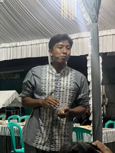 A medium shot captures a young Indonesian man standing under a white tent adorned with cascading drapes and string lights. He is wearing a short-sleeved grey shirt with a geometric pattern in white and black. His hands are positioned in front of him, one holding a small clear plastic bag. Behind him, tables are set with white tablecloths and teal chairs, suggesting a gathering or event. The scene appears to be outdoors, within the city of Wedarijaksa, Indonesia. The man's gaze is directed slightly upwards and to his left, with a neutral expression. The lighting suggests it is either dusk or nighttime.