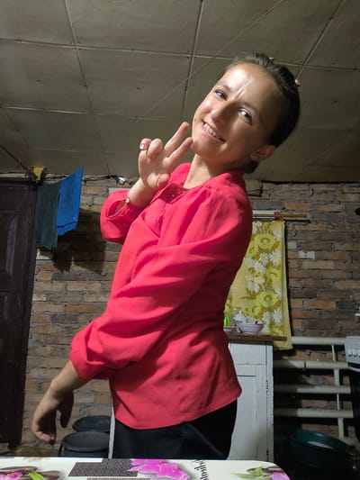 A young woman with dark hair pulled back is in a room with exposed brick walls and a textured ceiling. She is wearing a bright pink shirt and black pants, and she is smiling and holding up two fingers in a peace sign. Her pose suggests a casual and cheerful demeanor. The background shows a doorway with towels hanging on a rack, a yellow curtain with a floral pattern, and some kitchen items on a shelf. The lighting is dim, suggesting it might be evening or indoors with artificial light. The scene appears to be a humble living space, possibly a kitchen or utility area, in Anastasivka, Ukraine. There is no visible text in the image.