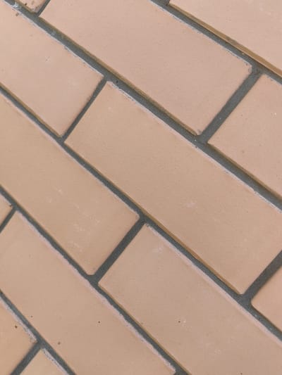 The image displays a close-up, angled view of a brick wall. The bricks are a light tan or pale orange color and are laid in a stretcher bond pattern, meaning they are staggered. The mortar between the bricks is a dark gray color. The surface of the bricks appears smooth, with some subtle variations in tone and texture, and a few small specks or imperfections are visible on some of the bricks. The lighting is even, and there are no people or other objects in the frame. The scene is a plain, textured surface, characteristic of an outdoor wall. This image could be of a wall located anywhere, and its specific location in Trudobelikovskiy, Russia cannot be determined from the visual information alone.