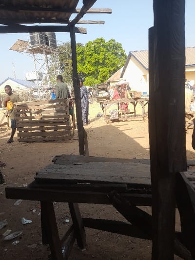 This image depicts an outdoor market scene in Wamba, Nigeria, under clear daylight conditions. The foreground features a dark, roughly constructed wooden table with a damaged plank tabletop, situated beneath an overhang of corrugated metal roofing supported by wooden beams. To the right, a vertical wooden post, part of the foreground structure, partially obscures the view.
The ground consists of dry, reddish-brown earth with visible debris, including white fragments (possibly plastic or paper).
In the midground, several individuals are present. On the left, a man wearing a light green shirt and dark shorts, appearing barefoot, holds a yellow container and faces towards the right. Behind him, another individual in a light green garment is partially visible. Centrally, a person in a patterned dark blue and white long garment stands, facing away or sideways, amidst other market activity.
Numerous improvised market stalls, constructed from wooden planks and pallets, are visible across the midground. Several distant wooden market tables display various goods, including fabrics and bags, likely under temporary coverings.
The background reveals a light yellow building with a brown sloped roof and a blue trim line on the right. To the left, a tall metal water tower supports two black water tanks. Green trees are interspersed throughout the background, with one large, prominent tree providing a focal point. The sky above is clear and blue. Shadows indicate bright, direct sunlight.