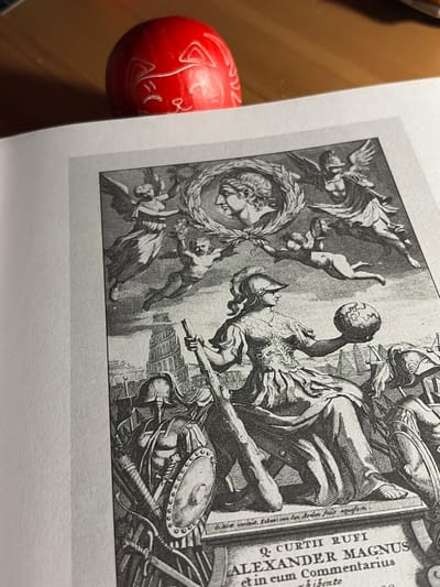 This is a close-up, black and white photograph of an etching or engraving, likely a book illustration or title page, with a red, carved object in the foreground. The etching depicts a dynamic, allegorical scene with multiple figures. In the center, a muscular figure, possibly a representation of Alexander the Great, is shown holding a globe aloft. Around him, other figures, some with wings, are in various states of movement, with one falling headfirst and another reaching out. The scene appears chaotic and dramatic, suggesting a narrative of triumph or upheaval. There are elements of classical attire, armor, and weaponry, including shields and swords. The background suggests an architectural or possibly a battlefield setting with detailed stonework and structures.
To the right of the etching, vertically oriented text is visible, reading: "Q. CURTII RUFI ALEXANDER MAGNUS et in eum Commentarius whibente". Below this, some fainter text is also present.
In the lower left corner, a bright red object is partially visible. It appears to be a carved stone or similar material, possibly an egg or a round seal, with etched symbols and markings. The object has a matte finish and its carved details stand out against the red surface. The overall lighting in the photograph is soft, highlighting the textures of both the print and the carved object. The setting appears to be indoors, with a neutral background that allows the subjects to be the primary focus.
