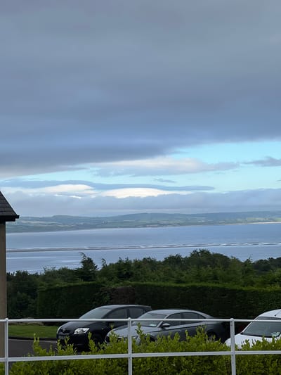 This image displays a scenic view of a body of water, likely a bay or estuary, with a distant coastline featuring rolling hills and vegetation. The sky is a mixture of dramatic clouds and patches of clear blue, suggesting dynamic weather. The water's surface is calm, reflecting the sky's colors.
In the foreground, a white railing separates the viewer from a neatly trimmed green hedge. Behind the hedge, several cars are parked. One dark-colored car is clearly visible, its headlights on, and part of its license plate is legible, reading "BZ67". Another white car is parked behind it. The parked cars and railing suggest a residential or parking area.
The time of day appears to be late afternoon or early evening, given the soft, diffused light and the quality of the sky. The location is Mountcharles, Ireland, and the overall atmosphere is peaceful and picturesque, capturing a tranquil moment by the water with hints of human presence in the foreground.