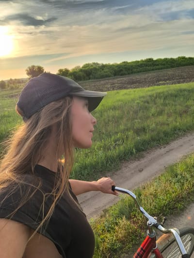 A young woman wearing a black t-shirt and a grey baseball cap is riding a red bicycle on a dirt road. The scene appears to be set in a rural area with fields and vegetation visible on both sides of the road. In the background, the sky shows a sunset or sunrise. The location cannot be precisely identified as Elets-Malanino, Russia without additional context.