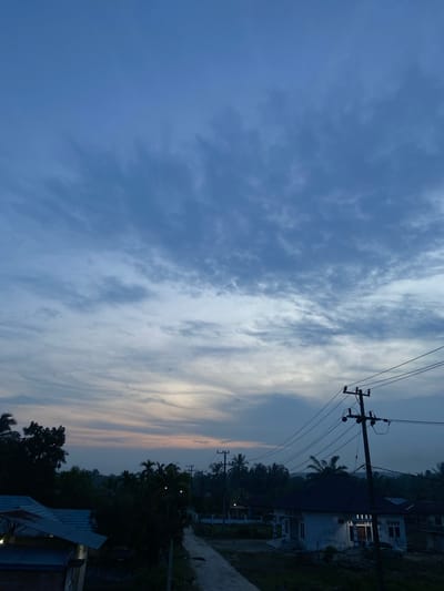 The image depicts a twilight scene in Candirejo, Indonesia. The sky is a blend of deep blues and lighter hues near the horizon, with scattered clouds. In the foreground, a dirt road leads into a village. To the right, a white house with a dark roof is illuminated by interior lights, and a utility pole with power lines stands nearby. On the left, the roof of a building is visible, and silhouetted trees dot the landscape. The overall atmosphere is quiet and serene, characteristic of early evening in a rural Indonesian setting.