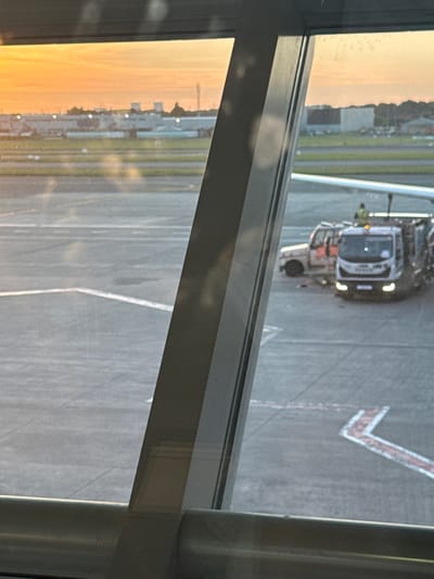 The image shows a view from inside an airport terminal at Brussels Airport in Zaventem, Belgium. The time appears to be sunset, with an orange sky visible in the background. In the foreground, there is a window frame. Outside the window, on the tarmac, are two vehicles, likely airport ground support equipment. One is a white truck with a person standing on top of it, and the other is a smaller white vehicle next to it. The tarmac is marked with white lines. In the distance, beyond the tarmac, there are buildings and industrial structures. The overall scene depicts a typical airport operational environment during twilight.