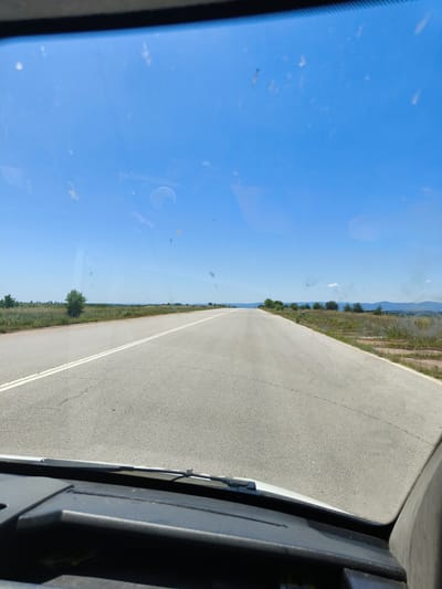 The image is a first-person perspective from inside a vehicle, looking through the windshield. The view is of an open, paved road stretching into the distance under a clear blue sky. The road has white dividing lines. On either side of the road, there are grassy fields with scattered trees and bushes. In the far distance, a mountain range is visible. The windshield has some smudges and debris on it, typical of everyday use. The lower part of the image shows the dashboard of the vehicle. The scene appears to be in a rural or semi-rural area, possibly on the outskirts of a town or village, with no other vehicles or people immediately visible. The sky is bright and sunny, suggesting daytime. Based on the information, this road view is likely in or near Mladinovo, Bulgaria, a location known for its rural landscapes.