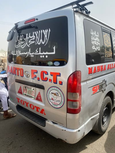 A silver Toyota Hiace commercial van is depicted from its rear three-quarter aspect, parked on an asphalt road in Gobirawa, Nigeria, under daylight conditions. The van features extensive religious and organizational decals. The rear window is darkly tinted, displaying large white Arabic calligraphy including "لا إله إلا الله محمد رسول الله" (There is no god but Allah, Muhammad is the messenger of Allah) and "لبيك يا رسول الله" (At your service, O Messenger of Allah). The rear-right side window also contains white and gold Arabic calligraphy.

The van's body displays "NARTO F.C.T." in large red and blue letters on the left rear, signifying the Nigerian Association of Road Transport Owners, Federal Capital Territory branch. A circular "NARTO FCT BRANCH" sticker is positioned below this text. The license plate, "KANO NSR-400ZL," is centrally mounted on the tailgate, flanked by three red triangular "CAUTION" stickers. The word "TOYOTA" is emblazoned in red on the lower part of the silver bumper, which exhibits scuffs and black marks on its lower right section. Further along the right side of the van, "MASHA ALLAH" and "SPORT" are visible in red and black lettering. A black roof rack is mounted atop the vehicle. The rear right wheel with a black rim is visible.

A person's lower legs and feet, clad in white attire and brown sandals, are partially visible on the left side of the frame, standing close to the van. The immediate environment consists of a paved road surface, with a hazy, bright background suggesting a dusty or distant urban landscape.