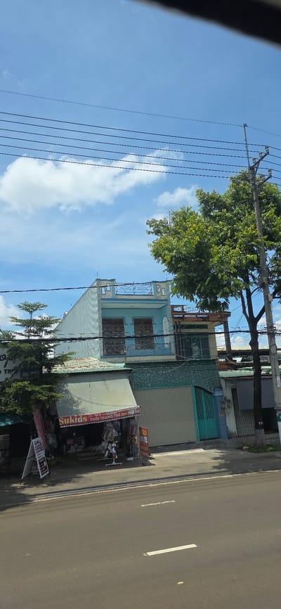 The image shows a street view in Thong Nhat Ward, Vietnam. In the foreground, there is a paved road with white lane markings. Beyond the road, a sidewalk is visible. On the sidewalk, there is a store with an awning and clothing racks displaying various garments. A sign above the store reads "Sukids THOI TRANG TRE EM". To the left of the store, a white A-frame sign stands on the sidewalk. Behind the store, a light blue building rises, featuring multiple stories, windows, and a balcony. A large green tree is situated to the right of the building, with its branches extending over the structure. Several power lines are strung across the sky, supported by utility poles. The sky is blue with scattered white clouds. The scene depicts a commercial area within a Vietnamese city.