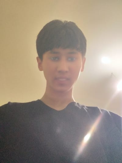 The image displays a low-angle portrait of a young person, likely a boy or young man, captured from a perspective looking upwards. The original image is rotated 90 degrees counter-clockwise.
The subject has short, dark hair, possibly with bangs, and is looking towards the camera. Their eyes are visible and dark, and their mouth is slightly open. The facial expression appears neutral or mildly inquisitive. They are wearing a dark-colored (likely black or dark grey) short-sleeved t-shirt.
The background is a plain, light-colored surface, resembling a ceiling or light-colored wall. Two bright light sources, possibly ceiling lights, are prominently visible in the upper part of the image, causing significant lens flare. One intense, multi-colored lens flare stretches diagonally across the person's right shoulder and chest, adding a bright streak to the composition. The overall lighting is bright and somewhat overexposed, particularly around the light sources and the flare, contributing to a slightly soft-focus and hazy quality to the image.
No specific activity is evident beyond posing for the photograph. The setting appears to be indoors. There is no visible text in the image.