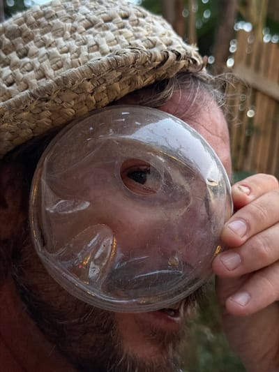 The image shows a close-up, profile view of a person wearing a woven hat, with their eye looking through a clear, round object held up to their face. The person has a beard and appears to be outdoors, with blurred greenery and wooden structures in the background. The lighting suggests it is daytime. The image does not contain any information that would allow for identification of the location within the city of Luang Prabang, Laos.